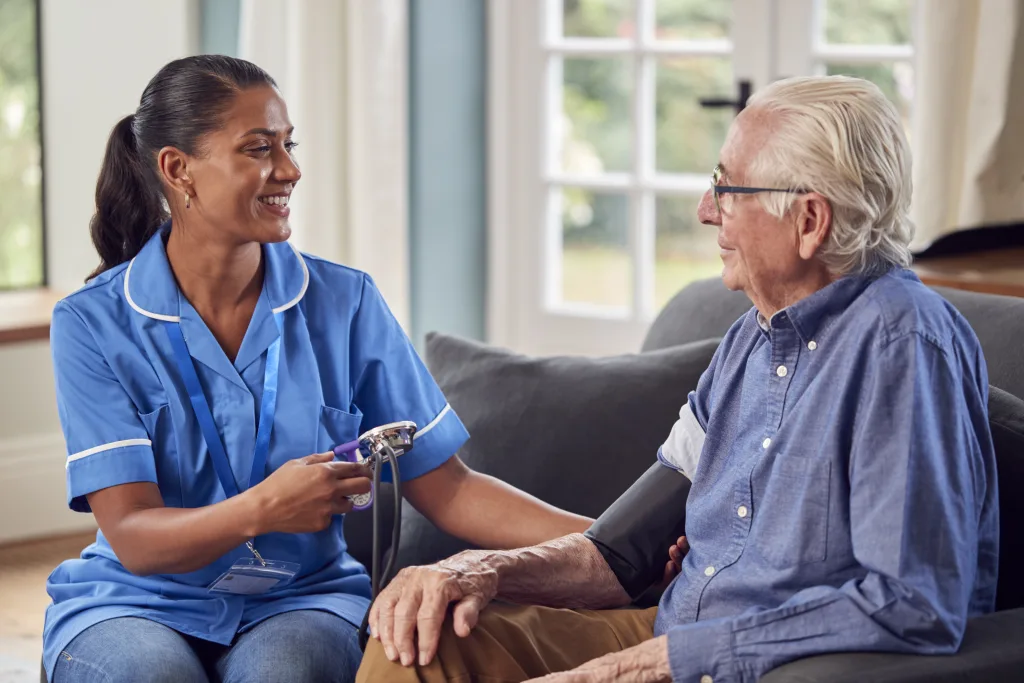Senior Man At Home In Lounge Having Blood Pressure Taken By Female Care Worker In Uniform